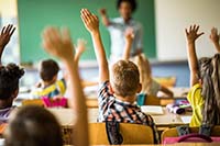 Photo of a teacher speaking to a classroom of students raising their hands